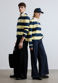Ριγωτά ράγκμπι μπλουζάκια σε ναυτικό μπλε και κίτρινο, συνδυασμένα με τζιν παντελόνια με φαρδιά παντελόνια σε σκούρο χρώμα. Παρουσιάζονται επίσης μια μαύρη τσάντα tote και ένα μαύρο καπελάκι με λογότυπο.
