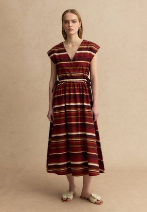 Mujer de pie, vestida con un vestido midi sin mangas de rayas en tonos granate y marrón, con la cintura ceñida y zapatos sin cordones de color crema.