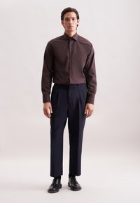 Camicia marrone a maniche lunghe con taschino, abbinata a pantaloni a pieghe blu navy e scarpe nere con lacci. Tessuto liscio e vestibilità su misura.