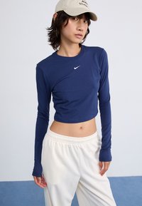 Tengerészkék, hosszú ujjú crop top kerek nyakkivágással és fehér Nike logóval, világosbarna, gumis derekú nadrággal párosítva. Simító, puha szövet textúra.