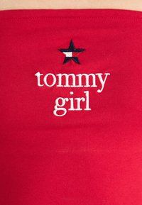 Rdeča bombažna tkanina z belim vezenim napisom "tommy girl" in mornarsko modro zvezdico nad napisom.