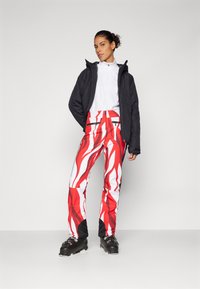 Schwarze isolierte Jacke, weißes Oberteil, rot-weiß gemusterte Skihosen mit schwarzen Akzenten und schwarze Skischuhe mit Schnallen.