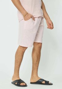 Shorts en lin rose clair avec poches latérales, associés à des sandales noires à brides croisées. Affichant une coupe décontractée et une texture respirante.