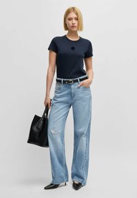 Marineblå bomulls-t-shirt med korta ärmar och broderad logotyp, tillsammans med ljusblå vida jeans med slitna detaljer, svart handväska och spetsiga klackar.