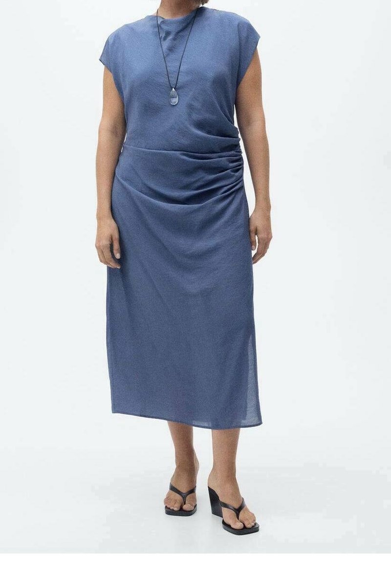Femme portant une robe midi bleu sans manches avec fronces sur les côtés et des sandales compensées noires, debout devant un fond blanc uni.