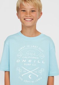 Šviesiai mėlynas trumpomis rankovėmis marškinėliai su balta grafika, vaizduojančia mešką, palmės ir persipynusias banglenčių lentas; pagaminti iš minkštos medvilnės medžiagos.