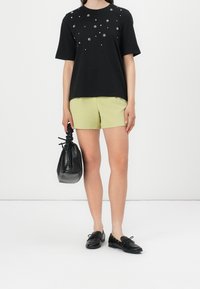 T-shirt noir à manches courtes avec des embellissements en clous, associé à un short vert clair. Tenant un sac noir texturé. Chaussures noires.