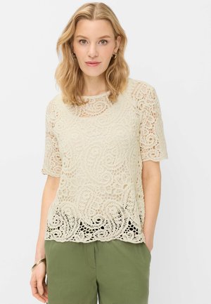 Kvinde med blondt hår iført en cremefarvet blonder-top med korte ærmer og olivengrønne bukser, stående med den ene hånd i lommen mod en hvid baggrund.