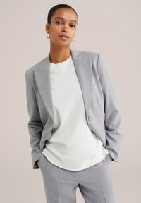Světle šedý přizpůsobený blazer s jedním knoflíkem, klopami a měkkou texturou, zkombinovaný s jednoduchou bílou košilí.