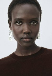 Modèle avec des cheveux courts et foncés portant de grandes boucles d'oreilles géométriques transparentes et un pull marron. L'arrière-plan est clair et neutre.