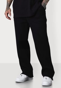 Schwarze Hose mit lockerer Passform, ausgestattet mit zwei Vorderseitentaschen und einer glatten Textur, kombiniert mit weißen Sneakers.