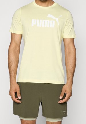 Hombre con una camiseta Puma de color amarillo claro y pantalones cortos Hyrox verde oliva con pantalones cortos de compresión beige debajo.