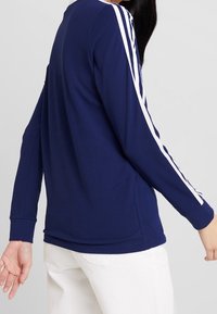 Tummansininen pitkähihainen paita, jossa on valkoiset raidat olkapäillä. Paita on valmistettu pehmeästä kankaasta, jossa on rento istuvuus ja pyöreä kaula-aukko.