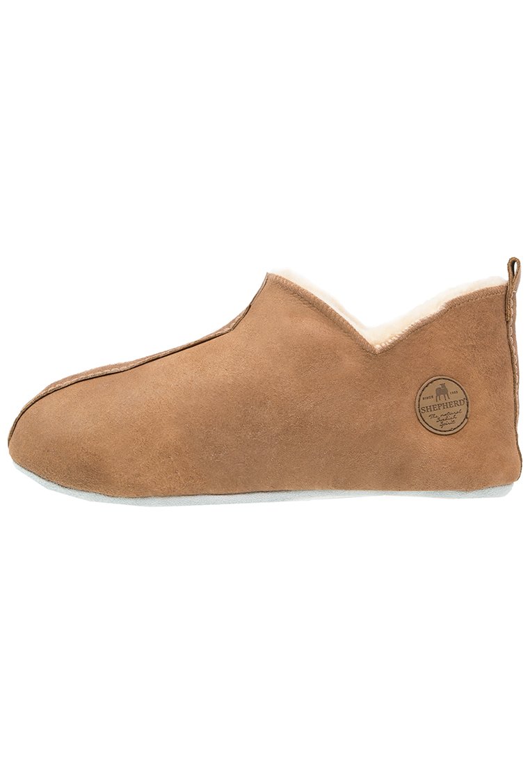 Chausson en suède marron avec un intérieur doux en blanc, semelle plate grise et une étiquette de marque circulaire sur le côté extérieur.
