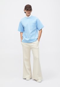 Camisa de mangas cortas azul claro con un bolsillo en el pecho, combinada con pantalones acampanados crema. El conjunto se completa con una gorra marrón y zapatos blancos.
