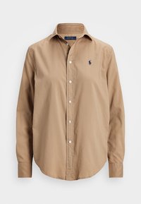 Ljusbrun skjorta med krage, tillverkad av ett mjukt tyg. Har en liten marinblå logotyp på vänster bröst och vita knappar. Långa ärmar.