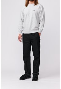 Homme portant une chemise à manches longues gris clair avec le logo "Makia", un pantalon noir et des chaussures noires, debout devant un fond clair uni.