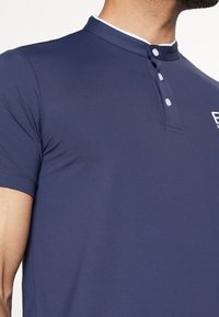 Námořnicky modré polo tričko s krátkými rukávy, kontrastním bílým límečkem, zapínáním na knoflíky a hladkou texturovanou látkou. Jemné logo značky na hrudi.