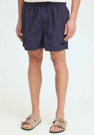 Mann trägt marineblaue Shorts mit Kordelzug und beige Sandalen mit doppeltem Riemen, steht vor einem schlichten hellen Hintergrund.