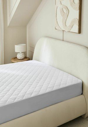 Einfaches Schlafzimmer mit gesteppter weißer Matratzenschoner auf einem cremefarben bezogenen Bett neben einem runden Holztisch mit dekorativer Lampe.