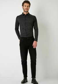 Schwarzes Hemd mit strukturiertem Liniendesign, kombiniert mit dunklen Jeans und schwarzen Stiefeletten. Das Modell steht vor einem neutralen Hintergrund.