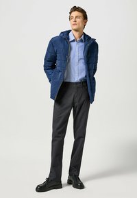 Marine gevoerd jack met capuchon, lichtblauwe blouse, donkergrijze broek en zwarte schoenen. Het jack heeft een ritssluiting aan de voorkant en zijzakken.