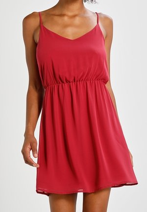 Femme portant une robe rouge sans manches avec des bretelles fines et une taille froncée, debout, les bras détendus, sur un fond clair.