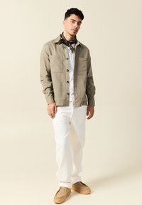 Lätt, beige jacka med stora fickor, bärs över en vit skjorta. Till det vita byxor och beige skor, accentuerat med en mönstrad scarf.