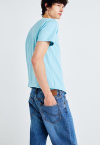 Young man in a light blue t-shirt and blue jeans with one hand in his back pocket, facing slightly sideways against a plain white background.
