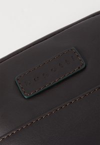 Dark brown leather bag with a glossy finish, featuring a rectangular embossed label with "bugatti" and contrasting brown stitching.