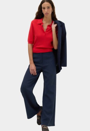 Kvinde iført en rød kortærmet skjorte med krave, marineblå vide bukser, sorte loafers, holder en marineblå blazer over skulderen.