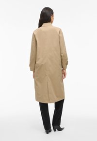 Donna in piedi, rivolta di spalle, indossa un lungo cappotto beige, pantaloni neri e stivali con tacco neri su uno sfondo bianco neutro.