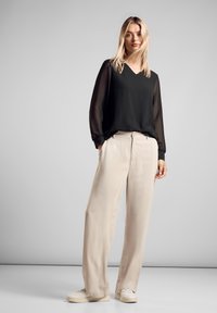 Blusa nera a maniche lunghe con scollatura a V e maniche trasparenti, abbinata a pantaloni beige a gamba larga e sneakers bianche, su uno sfondo neutro.