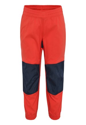 Peuter met krullend haar, beige campingthema T-shirt, rode en marineblauwe broek en witte sneakers, staand met duim in mond.