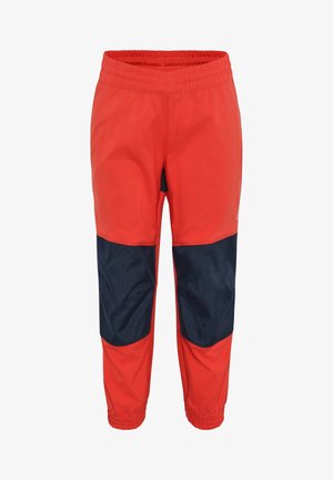 Kleinkind mit lockigem Haar, trägt beige Camping-T-Shirt, rote und marineblaue Hose und weiße Turnschuhe, steht mit Daumen im Mund.