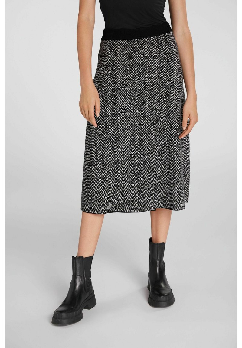 Femme portant un haut noir, une jupe midi à motif chevrons noir et blanc et des bottines noires épaisses, debout devant un fond uni.