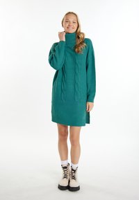 Abito maglione lavorato a maglia color teal con collo alto, caratterizzato da motivi a treccia e dettagli a coste; indossato con stivali alla caviglia color crema e calze bianche.