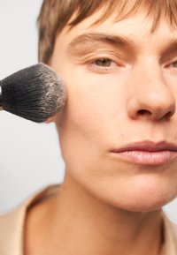 Un pennello per il trucco con setole nere morbide sta applicando della cipria sulla guancia di una persona con pelle liscia, toni neutri e trucco minimale.