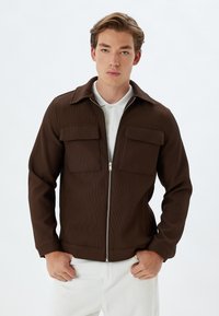 Chaqueta marrón de canalé con cremallera frontal, dos bolsillos en el pecho y cuello. Combinada con pantalones blancos, que muestran una textura de tela y un corte a medida.