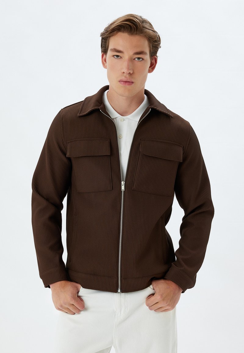 Chaqueta marrón de canalé con cremallera frontal, dos bolsillos en el pecho y cuello. Combinada con pantalones blancos, que muestran una textura de tela y un corte a medida.