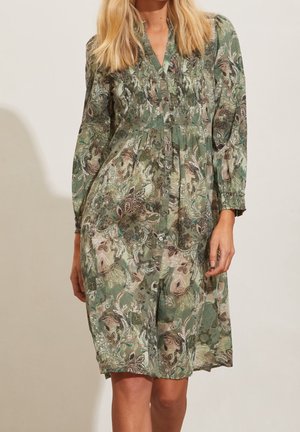 Femme portant une robe à manches longues, boutonnée sur le devant, avec un motif floral cachemire vert et beige et des détails smockés au niveau de la poitrine.