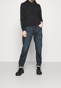 Felpa nera con cappuccio e lacci, con un piccolo logo rosso. Abbinata a jeans blu scuro, risvoltati alla caviglia, e stivali neri spessi.