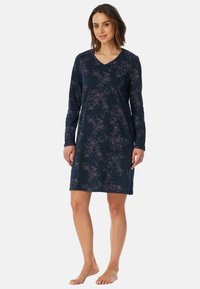 Donna in piedi a piedi nudi, con un vestito lungo a maniche, blu navy, con un motivo a contorno floreale rosa chiaro e scollo a V.