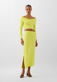 Completo due pezzi neon giallo che include un top aderente a maniche lunghe off-the-shoulder e una gonna abbinata con alta spacco, entrambi con una superficie liscia.
