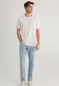 Helles rosa Baumwoll-T-Shirt mit Rundhalsausschnitt, kombiniert mit hellblauen Jeans. Das Model steht mit den Händen in den Taschen vor einem neutralen Hintergrund.