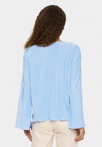 Blouse plissée bleu clair avec des manches longues et un col rond. Texture douce, poignets légèrement évasés et ourlet en ondulations.