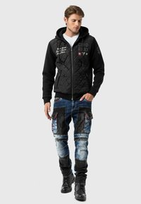 Gestepptes schwarzes Hoodie mit doppelten Texturen und Reißverschlüssen; kombiniert mit blauen und schwarzen Cargo-Jeans mit mehreren Taschen und Denim-Patches.