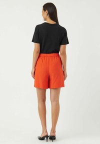 Musta lyhythihainen t-paita yhdistettynä oransseihin, teksturoidusti korkean vyötärön shortseihin. Mallilla on päällään mustat korkokengät, kuvassa näkyy takaa.