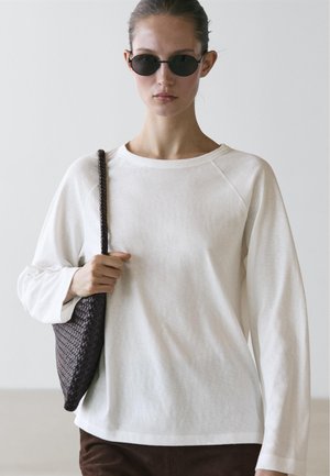 Frau mit schwarzen, runden Sonnenbrillen, weißem Langarmshirt, die eine dunkel geflochtene Schultertasche vor einem schlichten hellen Hintergrund hält.