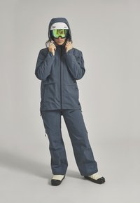 Ski jacket and pants in dark blue fabric, featuring multiple pockets, a zip closure, and a fitted hood. Black and white goggles with green lenses.